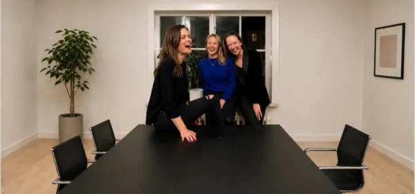 Three women laughing together around a dark meeting table in a bright room
