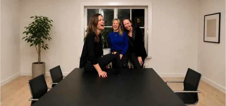 Three women laughing together around a dark meeting table in a bright room