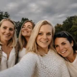 Four women smiling and taking a selfie together on the grass in a park