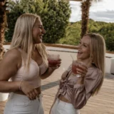 Two friends laughing together with drinks on a sunny terrace by the water