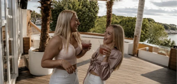 Two friends laughing together with drinks on a sunny terrace by the water
