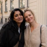 Two friends smiling with their heads close together on a quiet, tree-lined city street