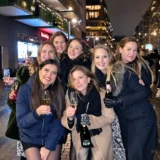 Seven women smiling together with glasses of bubbly on a city street at night