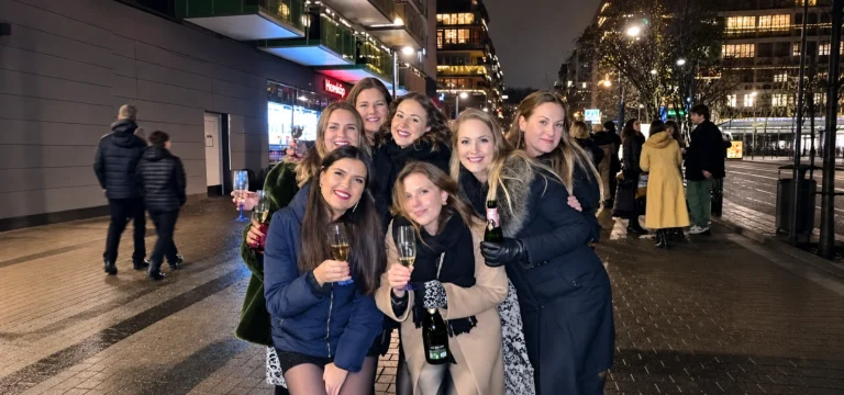 Seven women smiling together with glasses of bubbly on a city street at night