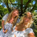 Two women in floral summer dresses and flower crowns taking a selfie in a sunny garden.