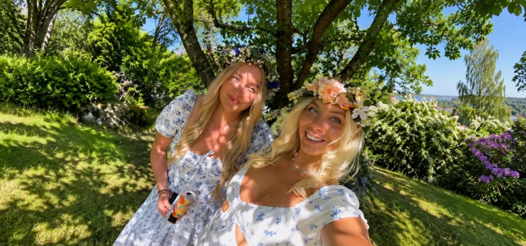 Two women in floral summer dresses and flower crowns taking a selfie in a sunny garden.