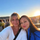 Two women taking a selfie together on a waterfront terrace at sunset.