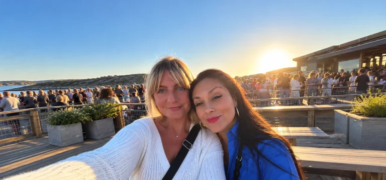 Two women taking a selfie together on a waterfront terrace at sunset.
