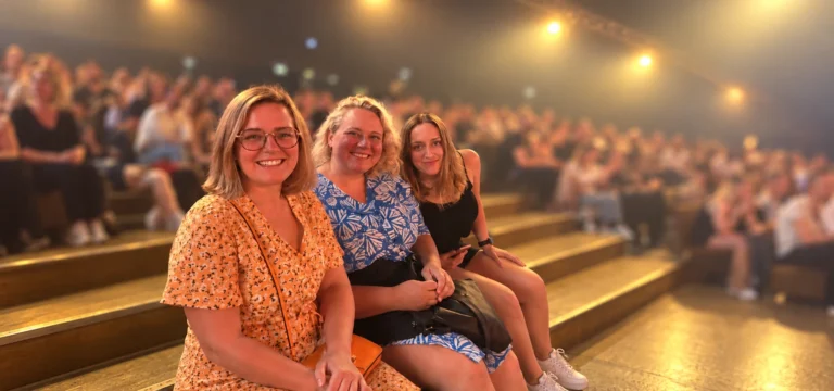 Three women sitting together on the steps of a theatre auditorium, smiling at the camera.