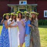 Five women in flower crowns raising their glasses together on a lawn in front of a red wooden house.